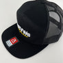 A black trucker-style cap with a flat brim, black mesh back panels, and white embroidered lettering on the front panel. A red and white Flexfit brand patch is visible on the brim.