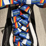 A close-up of a black low-top sneaker with white contrast stitching and white side panels, featuring colorful woven shoelaces with repeating blue, orange, yellow, and white text.