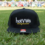 A black flat bill snapback trucker hat with a white embroidered 'hotvws' logo and yellow 'magazine' script on the front, featuring a red and white Richardson brand sticker on the brim, set in the grass with a Volkwagen beetles in the background